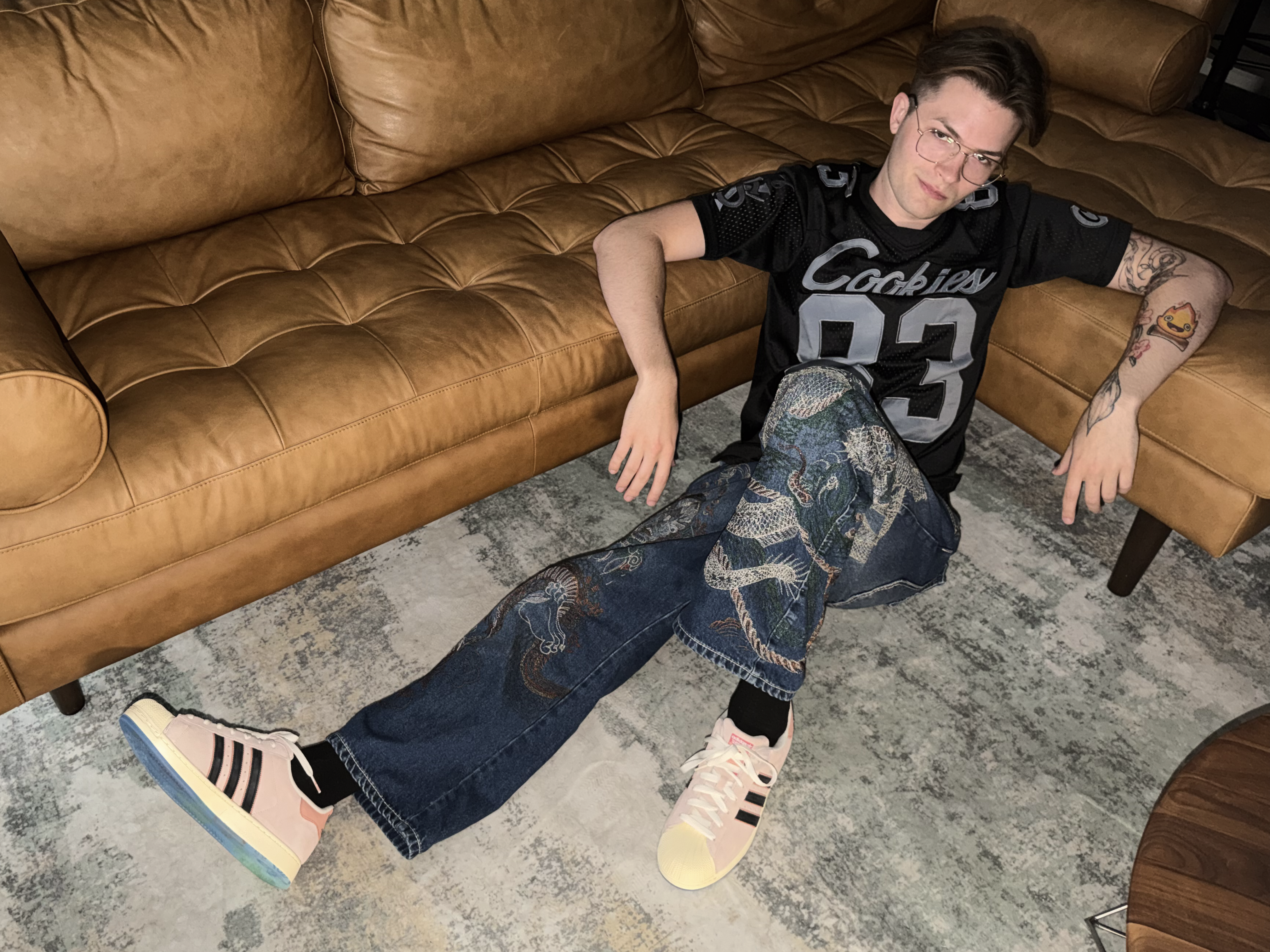 Nick sitting on a floor wearing a cool dragon embroidered denim jeans, Spongebob Squarepants shoes, and a black Cookies Jersey
